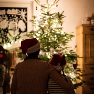 A parent hugs their child in front of a Christmas tree in a cozy dimly-lit room. They’re both wearing Santa hats.